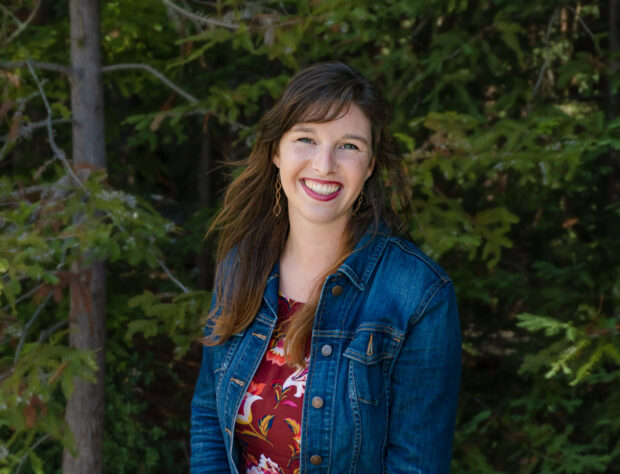 Anna Marie stands in front of Redwoods on UCSC's campus. She's grinning.