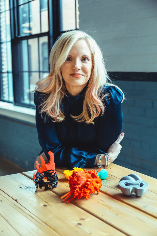 Kimberly Arcand sits at a table with six 3D prints of astronomical objects in front of her. These prints vary in color from orange, yellow, green, and gray. They are about the size of your hand.