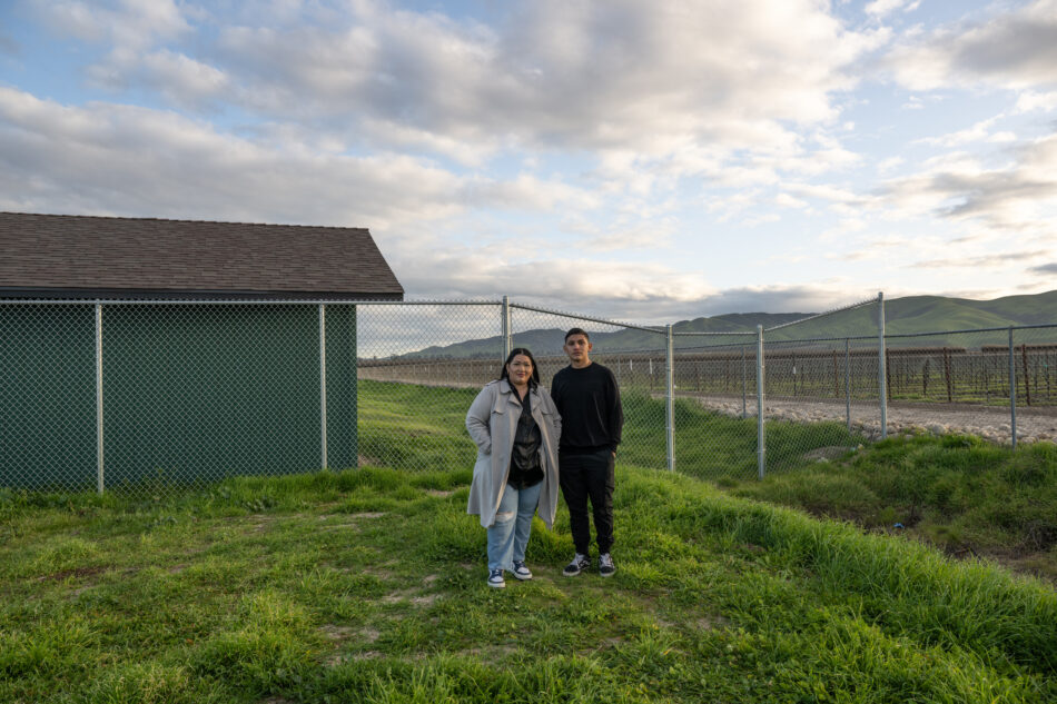 A woman and a young man stand in front of a fence which separates them from a green field. Behind them are rolling hills lit in afternoon light.