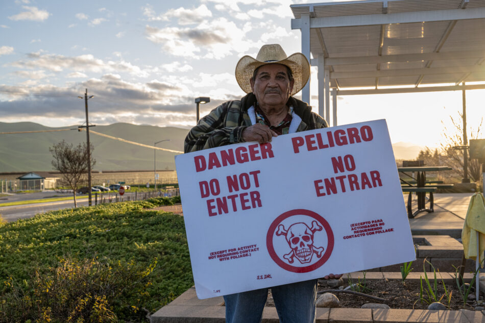 An older man in a cowboy had holds a large white sign which says "DANGER DO NOT ENTER/PELIGRO NO ENTRAR" in bold red letters around a red skull and crossbones.